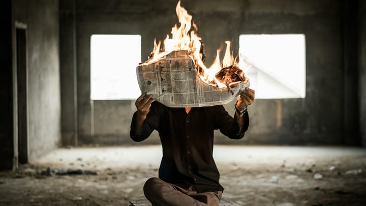 Person holding newspaper on fire, sat in a room that looks derelict with two blank windows behind.